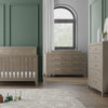 A modern nursery with green paneled walls features a wooden crib, dresser, and the Child Craft Universal Dressing Kit in Crescent Grey. Sunlight streams through white curtains, while toys and a rug add charm to the wood floor.
