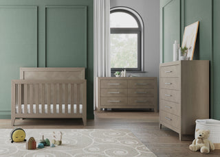 Modern nursery with green walls, a wooden crib, Child Craft's Kieran 5-Drawer Chest in Crescent Gray, a matching dresser, a window with a white curtain, and toys—plush sun, stacking rings, and teddy bear—on a white patterned rug.