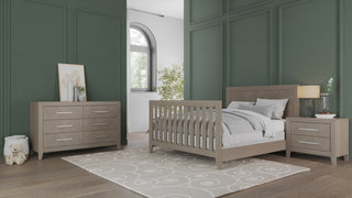 A modern bedroom with green paneled walls features the Child Craft Kieran 4-in-1 Convertible Crib in Crescent Gray, a matching nightstand and dresser, teddy bear, large window for natural light, and a beige patterned rug over wooden floors.