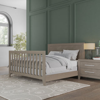 A modern bedroom features a wooden bed, gray and white bedding, a patterned rug, plant decor, and the Child Craft Kieran Nightstand in Crescent Gray with books and a lamp, set against green paneled walls by an arched window.