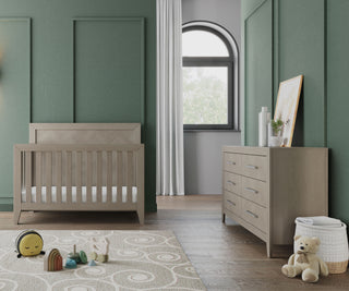 A modern nursery with green paneled walls, a Child Craft Kieran 4-in-1 Convertible Crib in Crescent Gray, matching dresser, toys on a patterned rug, teddy bear in a basket, and a large window with white curtains.