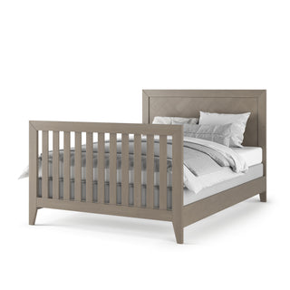 The Child Craft Kieran 4-in-1 Convertible Crib in Crescent Gray features a high headboard and matching footboard with vertical slats, evoking classic nursery design, and is styled with white and gray bedding and pillows.