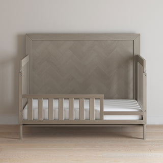 The Child Craft Toddler Guard Rail - Crescent Grey features a tall, patterned headboard, partial guard rail on one side, and a white mattress, set on a light wooden floor against a plain, light-colored wall.