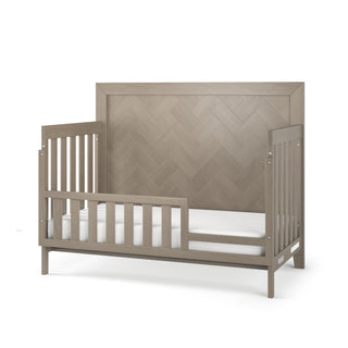 The Child Craft Kieran 4-in-1 Convertible Crib in Crescent Gray features slatted sides and a high herringbone-patterned headboard, shown without bedding—a stylish addition to any nursery furniture collection.