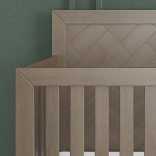 Close-up of the Child Craft Kieran 4-in-1 Convertible Crib in Crescent Gray, featuring a herringbone headboard, vertical side slats, and a green textured wall in the background.