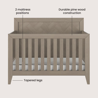 Front view of the Child Craft Kieran 4-in-1 Convertible Crib in Crescent Gray, showing vertical slats, chevron headboard, tapered legs, and text labels for 2 mattress positions and durable pine wood construction.