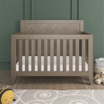 The Child Craft Kieran 4-in-1 Convertible Crib in Crescent Gray stands against a green paneled wall, with a plush bee, beige teddy bear, and woven basket on the wood floor, partly covered by a patterned rug.