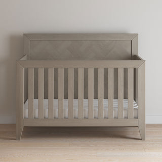 The Child Craft Kieran 4-in-1 Convertible Crib in Crescent Gray, featuring slatted sides and a solid geometric headboard, stands on a light wooden floor against a plain white wall.