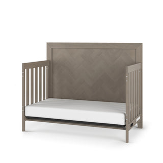 The Child Craft Kieran 4-in-1 Convertible Crib in Crescent Gray features a herringbone-patterned headboard, open sides with vertical slats, and a modern, neutral design that includes a white mattress.