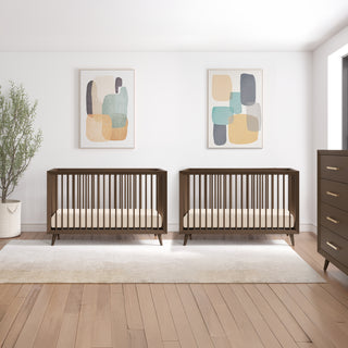 A modern nursery with two wooden cribs, abstract art on white walls, a potted plant, and the Child Craft Cranbrook 4 Drawer Chest in Toasted Chestnut, plus a light area rug on a wooden floor.