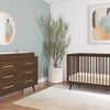 Modern nursery featuring a wooden crib, Child Craft dresser with the F01792 Universal Dressing Kit in Toasted Chestnut, round mirror, abstract wall art, potted plant, and beige rug on wooden floors with muted green and white walls.