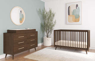 Modern nursery featuring a wooden crib, Child Craft dresser with the F01792 Universal Dressing Kit in Toasted Chestnut, round mirror, abstract wall art, potted plant, and beige rug on wooden floors with muted green and white walls.