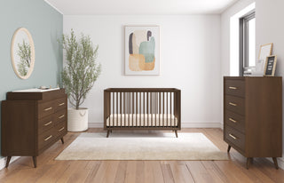 A modern nursery features a wooden crib, a Child Craft Cranbrook 4 Drawer Chest in Toasted Chestnut, abstract wall art, a potted plant, and a round mirror—all atop wood floors and a white rug in a bright, minimalist space.