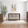 A modern nursery includes a wooden crib, Child Craft’s F01792 Universal Dressing Kit in Toasted Chestnut as a dresser/changing table topper, a light rug, potted plant, mirror, abstract wall art, and large window for natural sunlight.