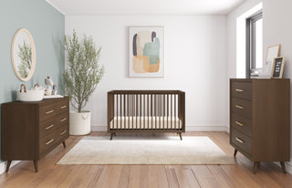 A modern nursery features a wooden crib, Child Craft Cranbrook 4 Drawer Chest in Toasted Chestnut, a round mirror, potted plant, abstract wall art on light walls, natural light from a window, and a beige rug on wooden floors.