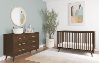 A modern nursery features a wooden crib, Child Craft Cranbrook Double Dresser in Toasted Chestnut with baby items, a round mirror, abstract art, a potted plant, soft green and white walls, wood floors, and a light area rug.