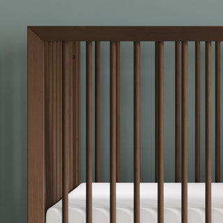 Close-up of the Child Craft Cranbrook 4-in-1 Convertible Crib in Toasted Chestnut, featuring vertical wooden slats and a white mattress against a muted green wall—perfect nursery furniture for a stylish space.