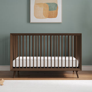 The Child Craft Cranbrook 4-in-1 Convertible Crib in Toasted Chestnut with vertical slats and a white mattress stands against a green wall, framed by modern nursery decor including wood floors and a white rug.