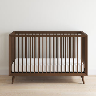 The Child Craft Cranbrook 4-in-1 Convertible Crib in Toasted Chestnut features sleek vertical slats and a white mattress, resting on a light wood floor against a white wall—ideal stylish nursery furniture.
