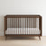 The Child Craft Cranbrook 4-in-1 Convertible Crib in Toasted Chestnut features sleek vertical slats and a white mattress, resting on a light wood floor against a white wall—ideal stylish nursery furniture.