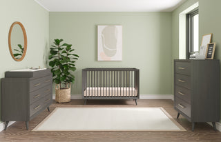 A modern nursery with sage green walls, Child Craft's Cranbrook 4-in-1 Convertible Crib in Lunar Gray, coordinated gray nursery furniture and changing table, a large potted plant, abstract art, and a white rug on wood flooring.