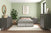 A modern bedroom with sage green walls, a double bed with gray and beige bedding, a Child Craft Cranbrook Double Dresser in Lunar Gray, another mid-century dresser, a nightstand, a potted plant, abstract wall art, and a round mirror above a dresser.