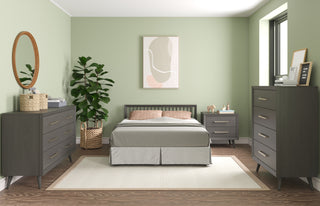 Modern bedroom with sage green walls, neutral bedding, dark wood dressers, a nightstand, and a large plant. The Child Craft Cranbrook 4-in-1 Convertible Crib in Lunar Gray adds mid-century flair, complemented by abstract art and abundant natural light.
