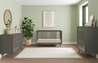 A modern nursery with sage green walls, a gray crib, Child Craft's Cranbrook 4 Drawer Chest in Lunar Gray, a matching dresser, a large green plant in a basket, a circular mirror, minimalist wall art, and a light rug on wood floors.