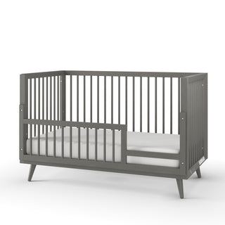 The Child Craft Cranbrook 4-in-1 Convertible Crib in Lunar Gray features slatted sides, angled legs, and a partial safety rail, bringing modern mid-century style to your nursery. Shown against a white background.