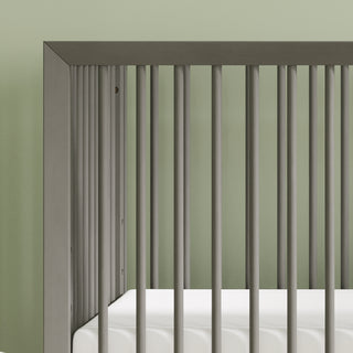 A close-up of the Child Craft Cranbrook 4-in-1 Convertible Crib in Lunar Gray with vertical slats and a white mattress, set against a light green wall—an ideal nursery furniture choice.