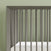 A close-up of the Child Craft Cranbrook 4-in-1 Convertible Crib in Lunar Gray with vertical slats and a white mattress, set against a light green wall—an ideal nursery furniture choice.