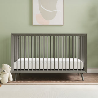 The Child Craft Cranbrook 4-in-1 Convertible Crib in Lunar Gray, paired with a white mattress, stands against a light green wall. A teddy bear rests beside it on wooden floors with a white rug and abstract art above the crib.