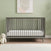 The Child Craft Cranbrook 4-in-1 Convertible Crib in Lunar Gray, paired with a white mattress, stands against a light green wall. A teddy bear rests beside it on wooden floors with a white rug and abstract art above the crib.