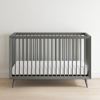 The Child Craft Cranbrook 4-in-1 Convertible Crib in Lunar Gray, featuring vertical slats and a white mattress, is placed on a light wood floor against a plain white wall.