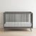 The Child Craft Cranbrook 4-in-1 Convertible Crib in Lunar Gray, featuring vertical slats and a white mattress, is placed on a light wood floor against a plain white wall.