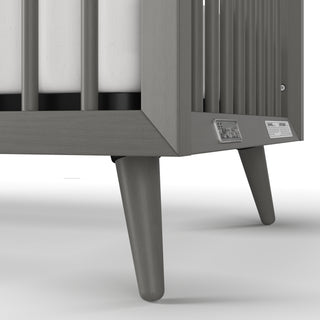 Close-up of the Child Craft Cranbrook 4-in-1 Convertible Crib in Lunar Gray, featuring tapered legs, vertical slats, and visible mattress. Two metal safety labels are attached to the frame—ideal nursery furniture for a stylish space.