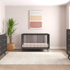 A modern nursery featuring a black crib, black dressers with the Child Craft Universal Changing Table Topper in Ebony (F01792), a beige rug, round mirror, potted plant, and neutral wall art. Light walls and wood floors with a right-side window brighten the space.