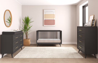 A modern nursery featuring a black crib, black dressers with the Child Craft Universal Changing Table Topper in Ebony (F01792), a beige rug, round mirror, potted plant, and neutral wall art. Light walls and wood floors with a right-side window brighten the space.