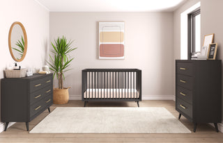 A modern nursery with a black crib, Child Craft Cranbrook Double Dresser in Ebony, a matching chest, abstract wall art, a potted plant, a round mirror, and a light cream rug on wooden floors bathed in soft natural light.