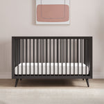 The Child Craft Cranbrook 4-in-1 Convertible Crib in Ebony with a white mattress stands against a light wall, topped by a minimalist pink and beige abstract painting, while a beige rug warms the wooden floor beneath the nursery setup.
