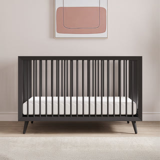 The Child Craft Cranbrook 4-in-1 Convertible Crib in Ebony with a white mattress stands against a light wall, topped by a minimalist pink and beige abstract painting, while a beige rug warms the wooden floor beneath the nursery setup.