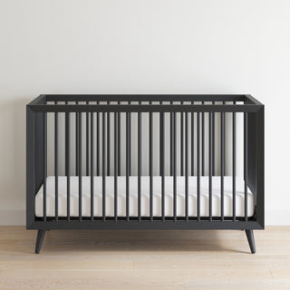 The Child Craft Cranbrook 4-in-1 Convertible Crib in Ebony, featuring vertical slats and a white mattress, adds stylish versatility to your nursery with its modern design, set on a light wooden floor against a plain white wall.