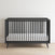 The Child Craft Cranbrook 4-in-1 Convertible Crib in Ebony, featuring vertical slats and a white mattress, adds stylish versatility to your nursery with its modern design, set on a light wooden floor against a plain white wall.