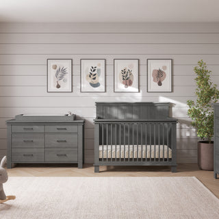 A modern nursery with gray furniture, including a crib, Child Craft dresser, and the F01792 Universal Changing Table Topper in Midnight Gray. Décor features a rocking bunny, plant, framed art on white shiplap walls, and sunlight on a beige rug.