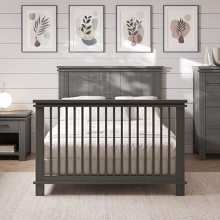A Child Craft F06424 Full-Size Bed with Midnight Grey rails is centered in a nursery, flanked by matching furniture. Framed abstract art hangs on a white shiplap wall above, softly illuminated by lamplight.