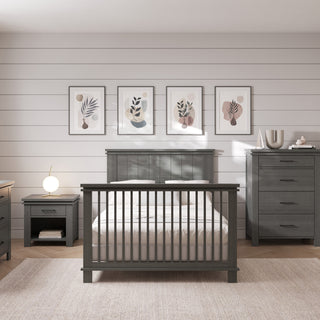 A modern gray-toned bedroom features Child Craft’s F06424 Full-Size Bed Rails in Midnight Grey, paired with a nightstand and dresser, white bedding, a round lamp, four minimalist botanical prints on shiplap, and a light rug over wood floors.