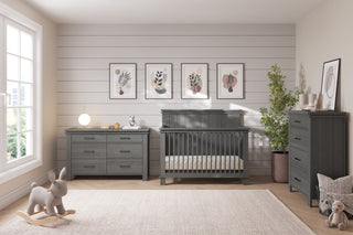 A modern nursery features gray wooden furniture, including the Child Craft Denman Double Dresser in Midnight Gray, a crib, and chest of drawers. Art prints hang on shiplap walls; a plush bunny rocker and potted plant complete the look.