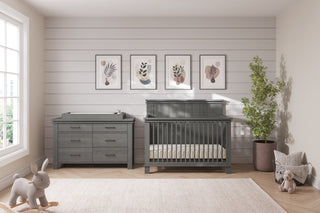 A neutral nursery featuring a gray crib, Child Craft Denman Double Dresser in Midnight Gray, potted plant, plush bunny toy, botanical wall prints, and soft natural light from a large window.