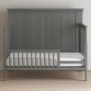 A Child Craft F09503 Toddler Guard Rail in Midnight Gray is installed on a toddler bed with slatted sides, paired with white bedding and mattress, set on a light wood floor against an off-white wall.