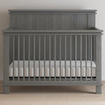 The Child Craft Denman 4-in-1 Convertible Crib in Midnight Gray features vertical slats, a high solid back, and a white mattress, shown on a light wood floor against a plain white wall.